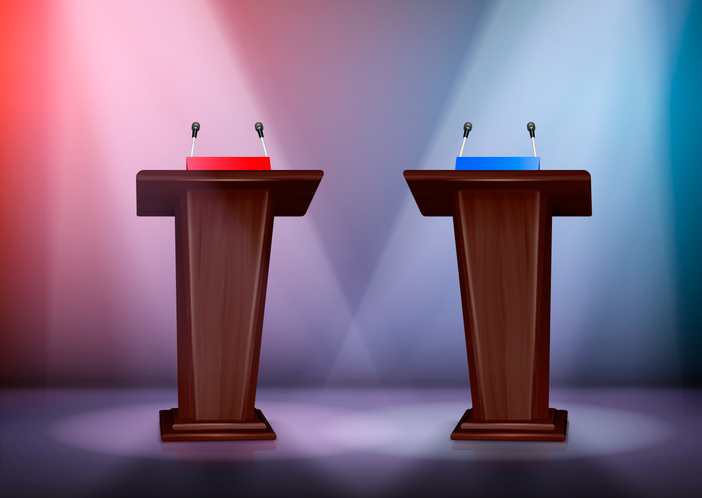 Two podiums with microphones are on a stage, the podium on the right is under red spotlights while the one on the left is under blue lights.