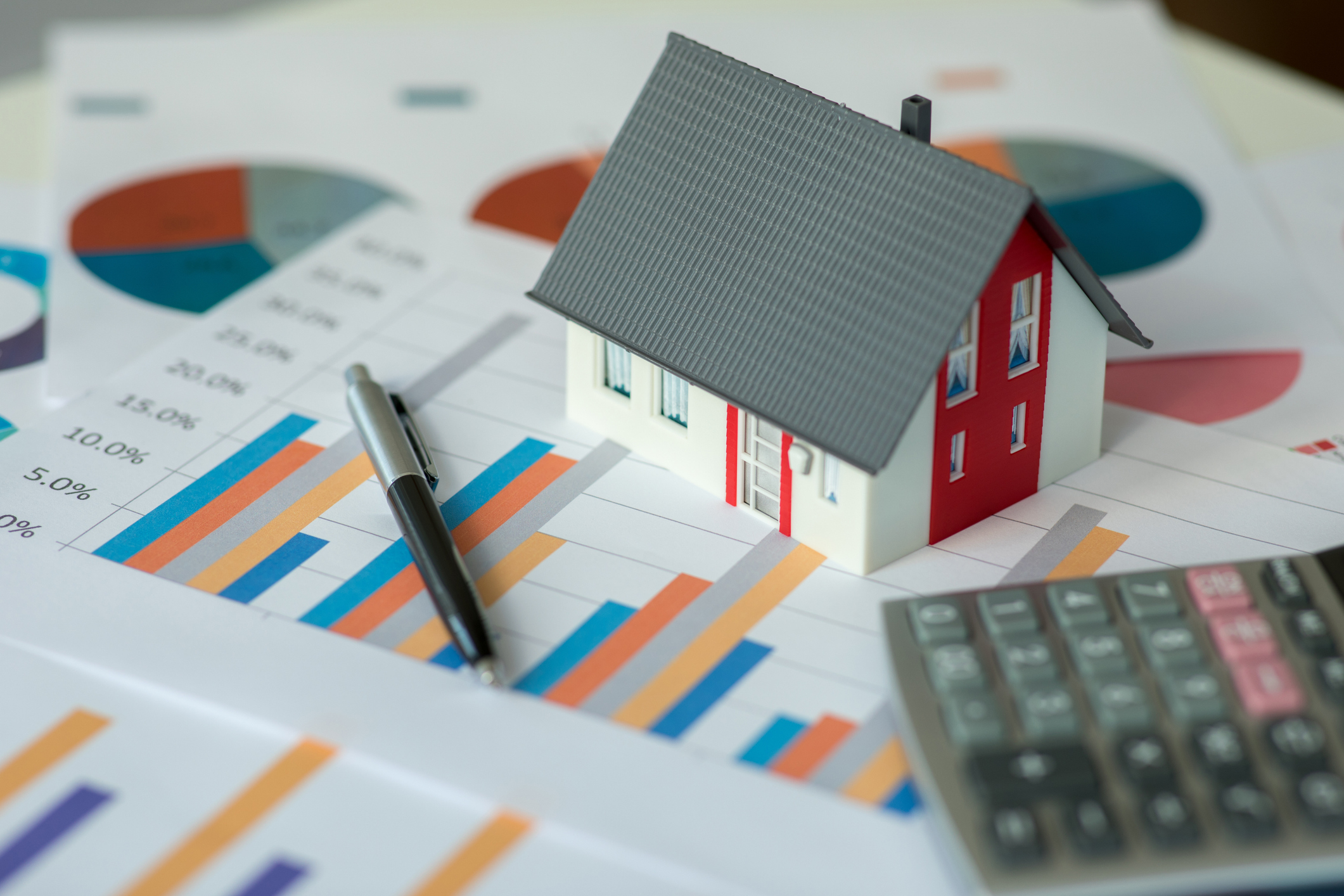 papers on desk with graphs on them, with a calculator and a small toy house on top of the papers