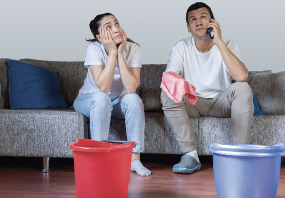 two people sitting on a couch looking up at the ceiling with buckets on the floor