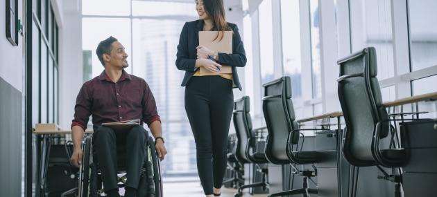 man in wheelchair walking down office hallway with woman holding file folder