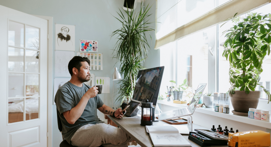 man sitting at desk in front of computer with a coffee, looking out the window pensively
