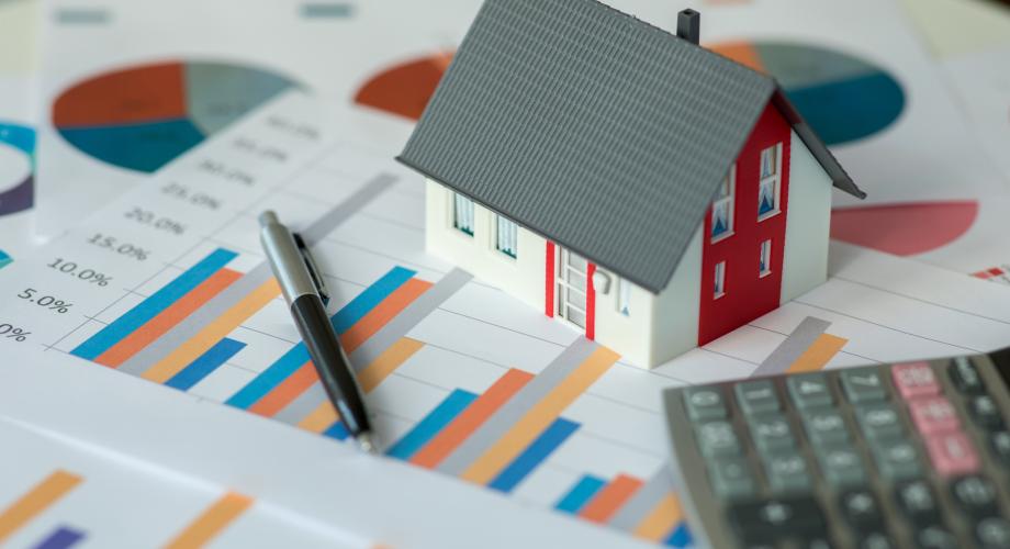 papers on desk with graphs on them, with a calculator and a small toy house on top of the papers