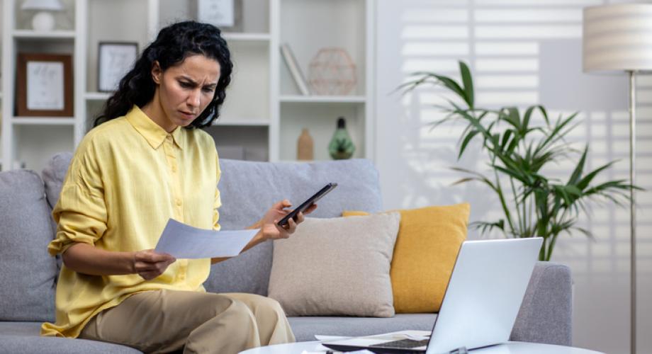 Dissatisfied and disappointed woman behind paper work sitting on sofa at home, Hispanic woman holding utility bills, creditor checks, paying upset using phone app.