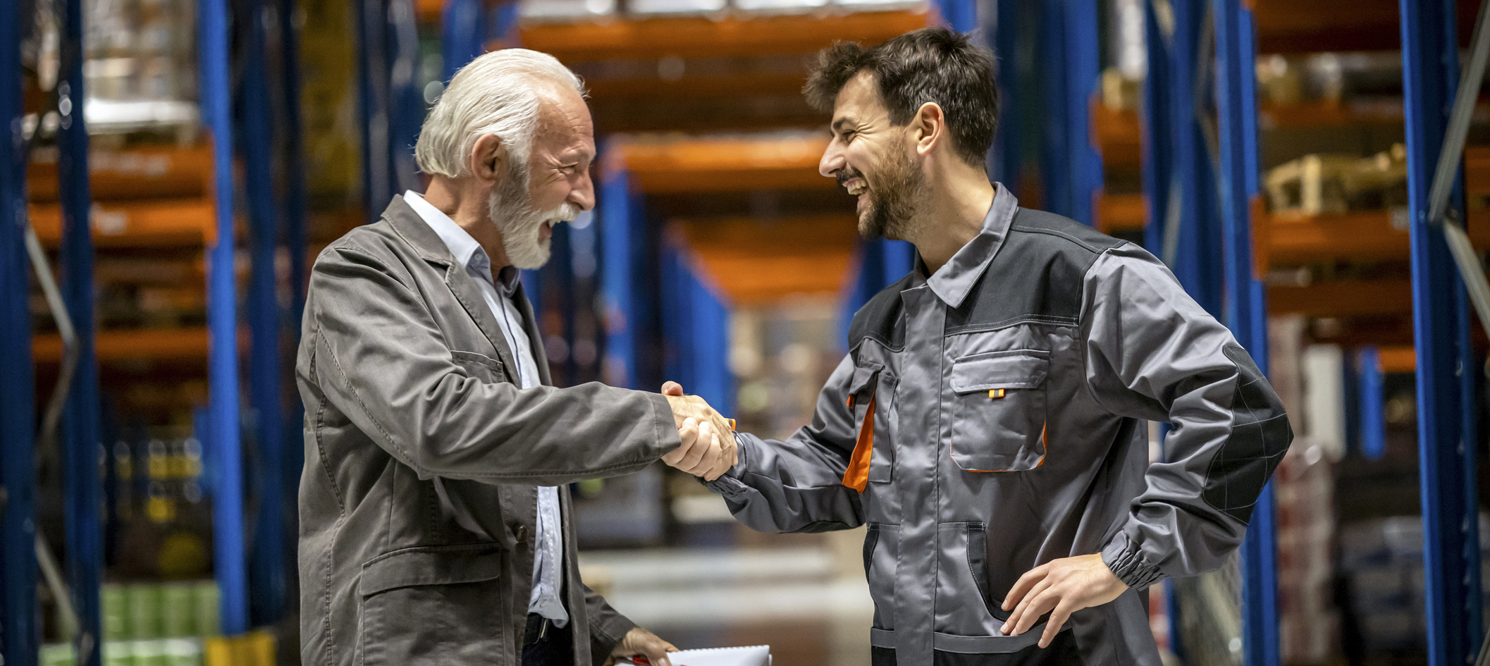 an older man and a younger man shaking hands and smiling in a warehouse